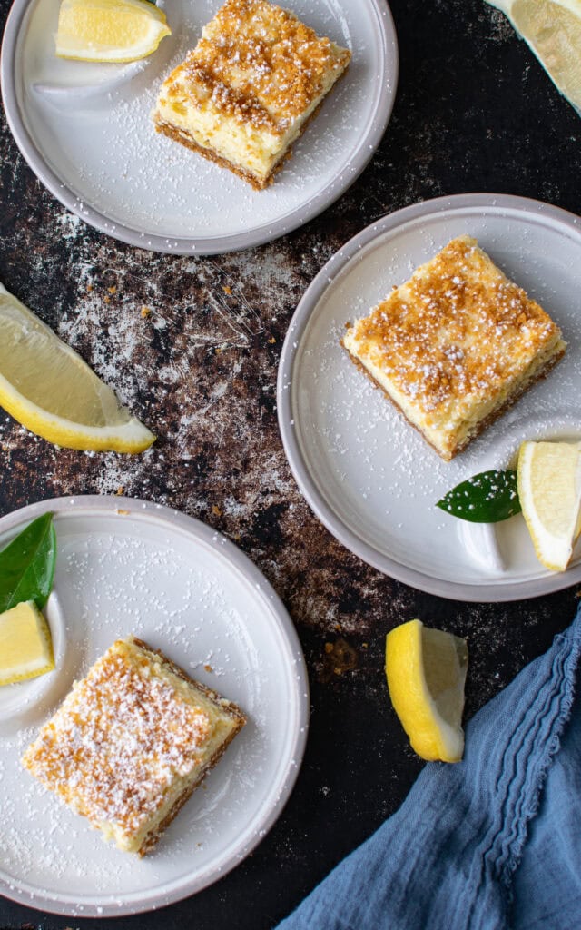 Looking down on two plates with graham cracker topped lemon squares on round plates with a wedge of lemon on each plate.