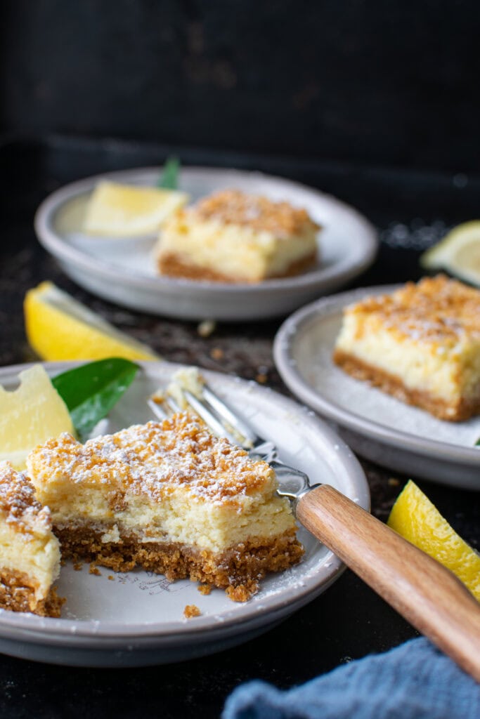 3 Lemon squares with a graham cracker crumble on plates.  One has a bit taken from it.