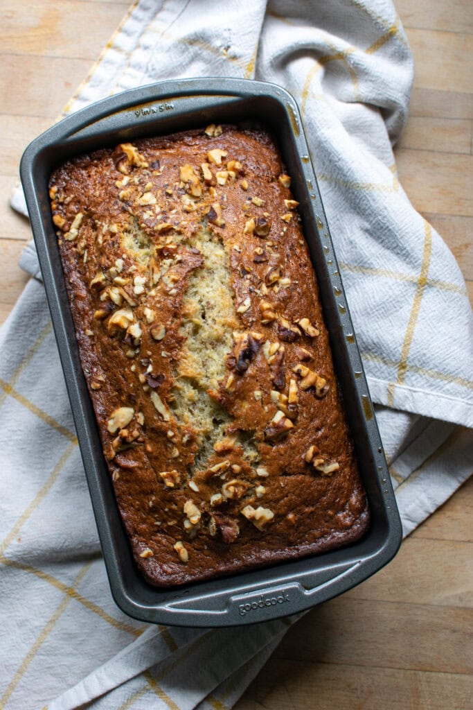 A loaf of banana rum bread topped with walnuts just out of the oven and resting on a white and yellow  kitchen towel.