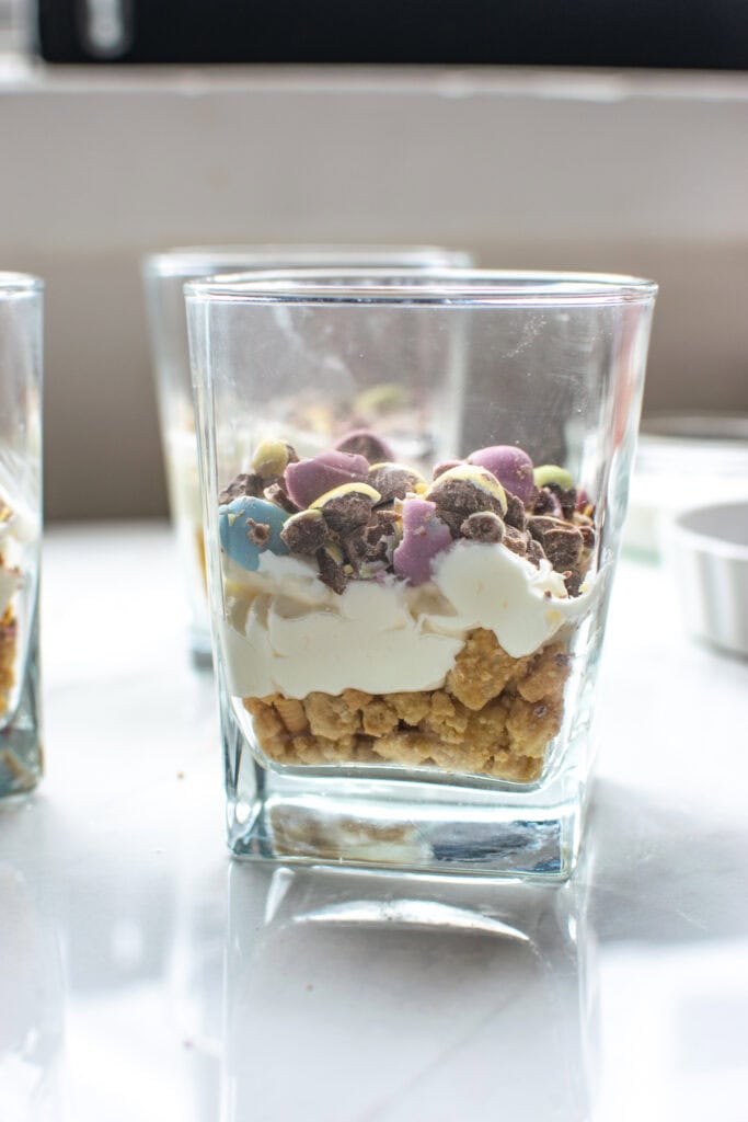 The side view of a dessert glass with a layer of cookie crumbs, a layer of cream cheese mixture, and a layer of crushed chocolate Easter mini eggs.