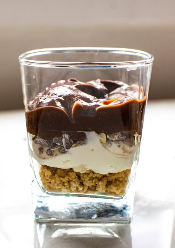 A dessert glass with a layer of vanilla cookie crumbs, a layer of cream cheese mixture, a layer of crushed chocolate mini eggs, and a layer of chocolate pudding.