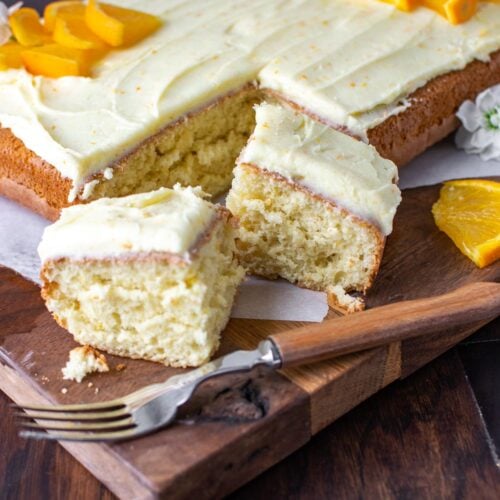 An orange sheet cake, frosted with orange butter cream and fresh oranges. It has two slices cut out of one corner.