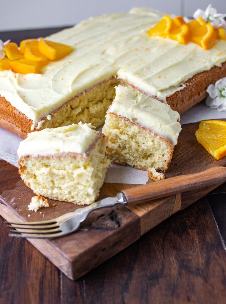 An orange sheet cake, frosted with orange butter cream and fresh oranges.  It has two slices cut out of one corner.